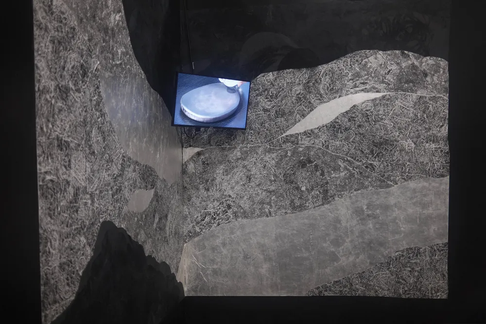 Installation view of Where Mountains Meet, a wall-scale collage of stone rubbings, with a small video monitor